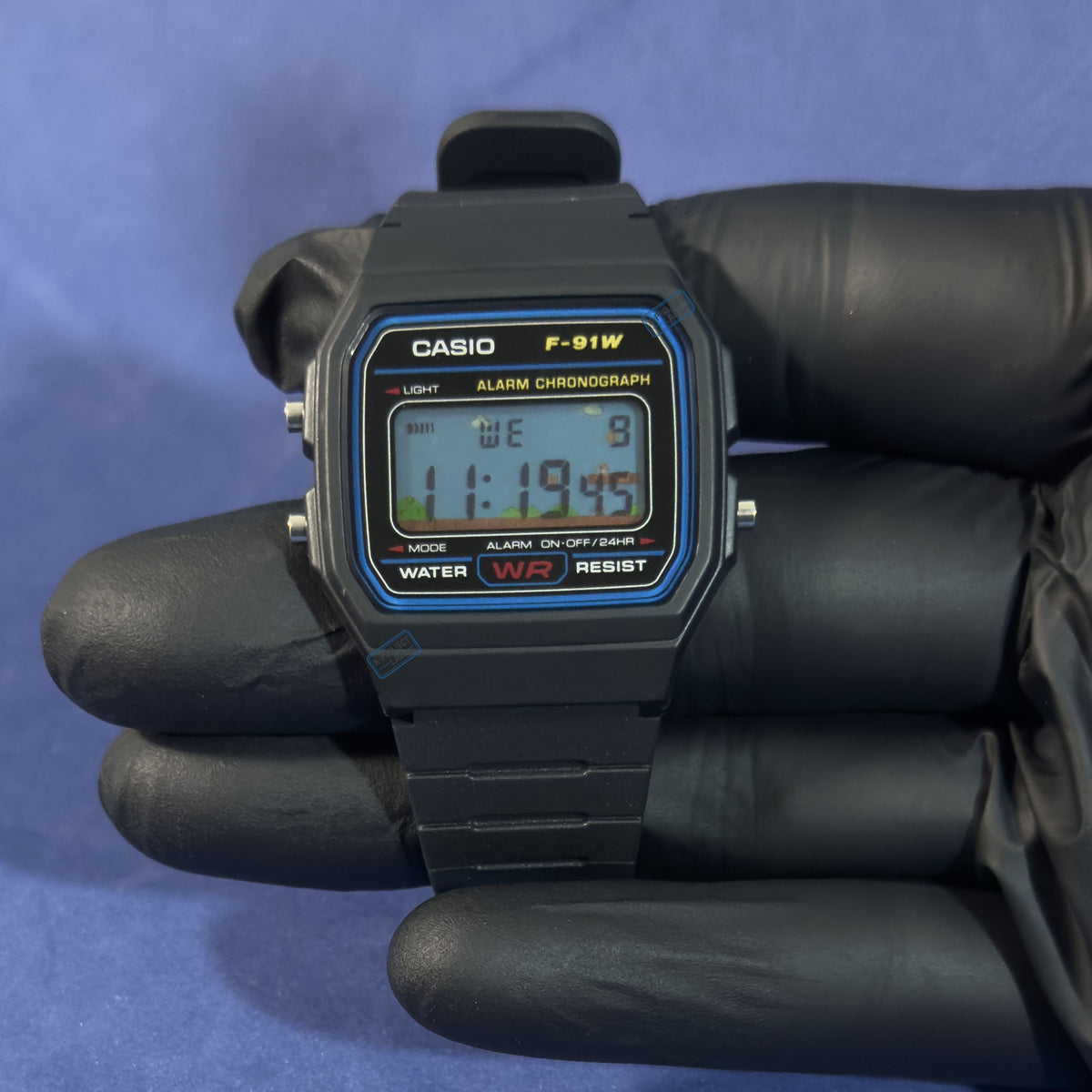 K-Project Labin Casio F-91W Pixel Kingdom mustana hartsina, mustassa hansikkaassa kädessä, pikselöity näyttö