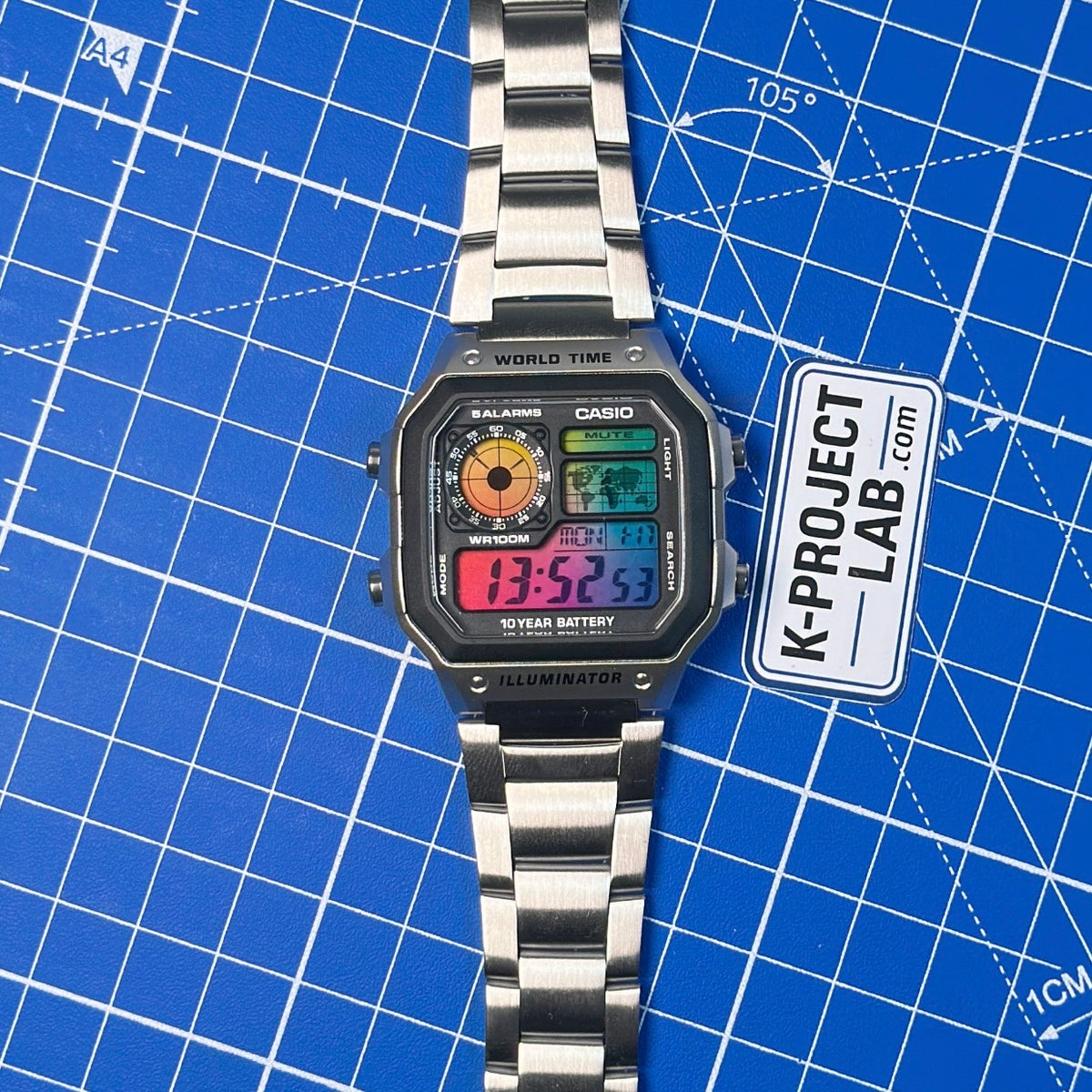 Casio AE-1200 Amber Signal in Stahl von vorne auf blauer Fläche, buntes Display, von K-Project Lab