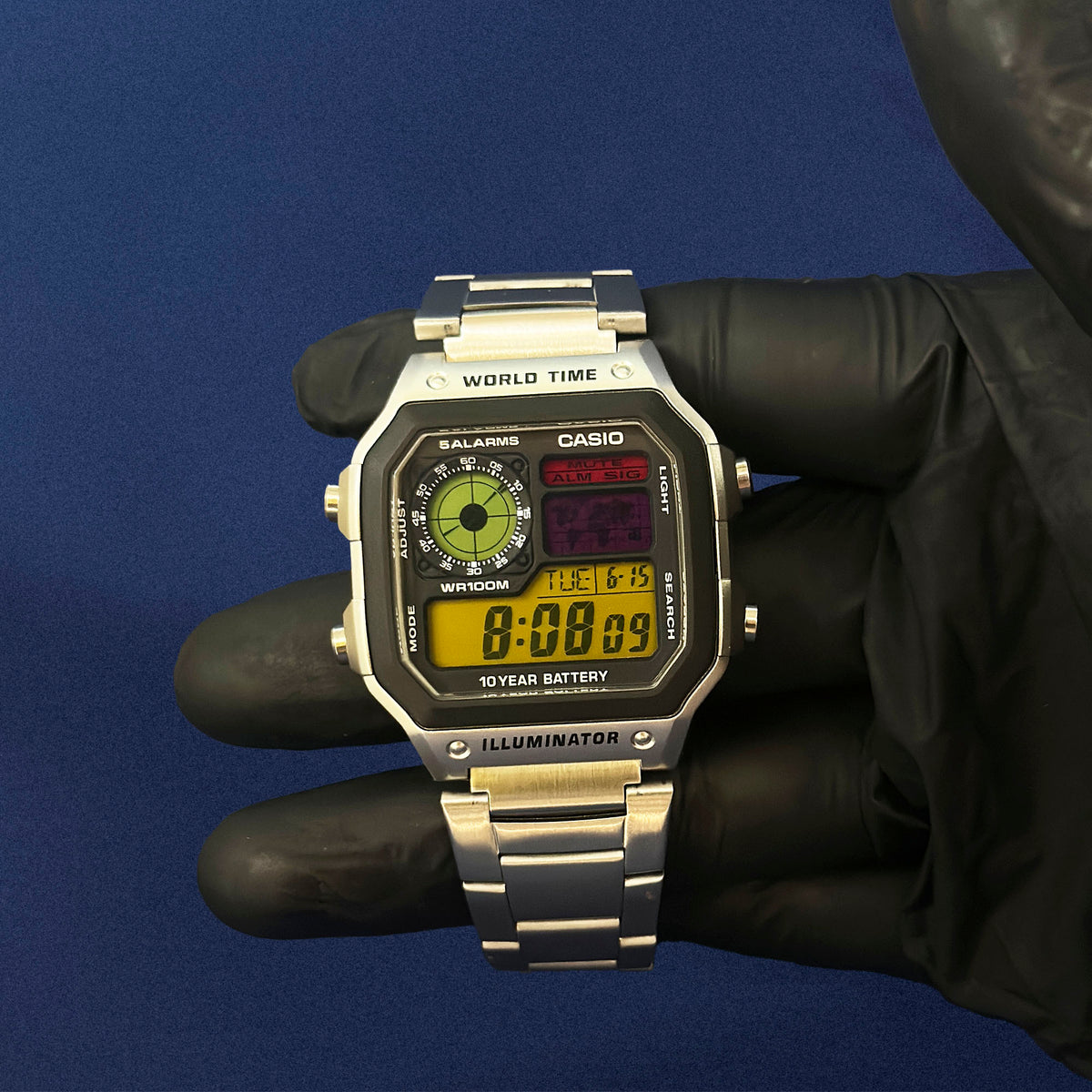 Casio AE-1200 Retro Frequency in Stahl, in schwarz behandschuhter Hand vor blauem Hintergrund, von K-Project Lab